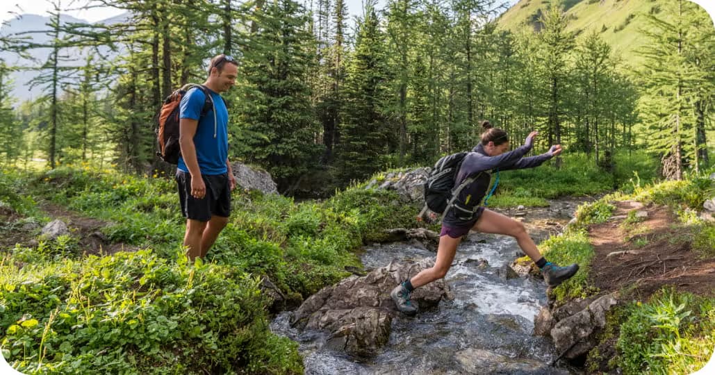 Two hikers jumping over a small creek one at a time in the middle of a forest